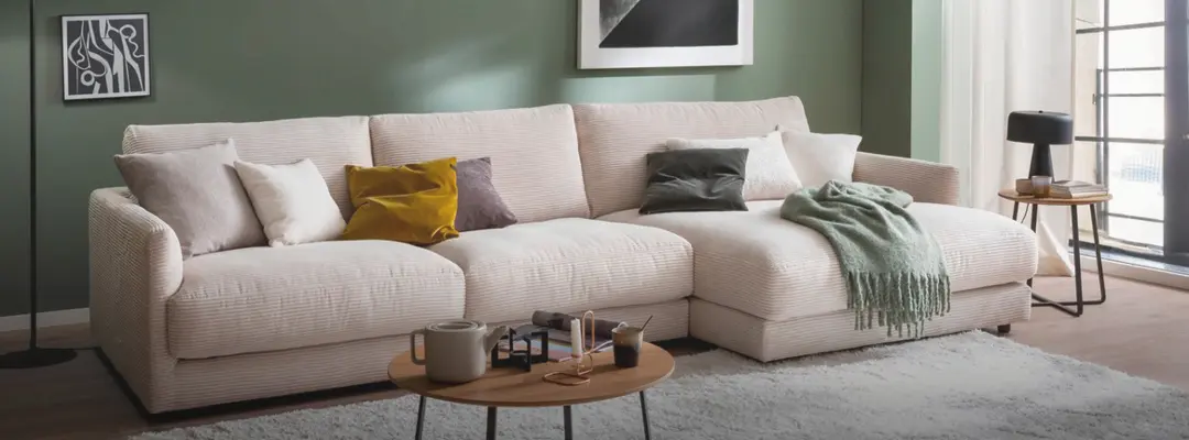 Helles Ecksofa mit Kissen, Couchtisch und Teppich vor grüner Wand und Fenster